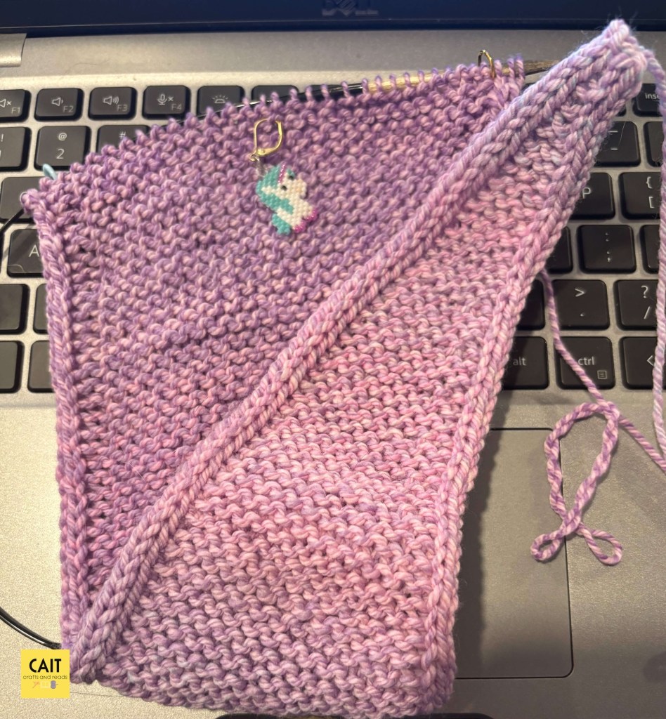 A hand knit scarf in progress in a marled pink, blue, and purple yarn. There is a beaded unicorn stitch marker. The project is on an open laptop.