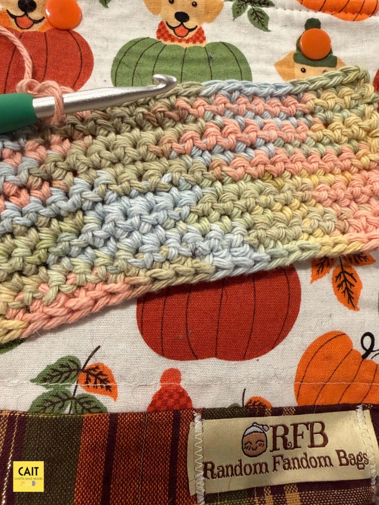 A crochet work-in-progress in single crochet stitch. The yarn is a mix of pastel shades. The project lays on top of a fall-themed project bag with pumpkins and golden retrievers.